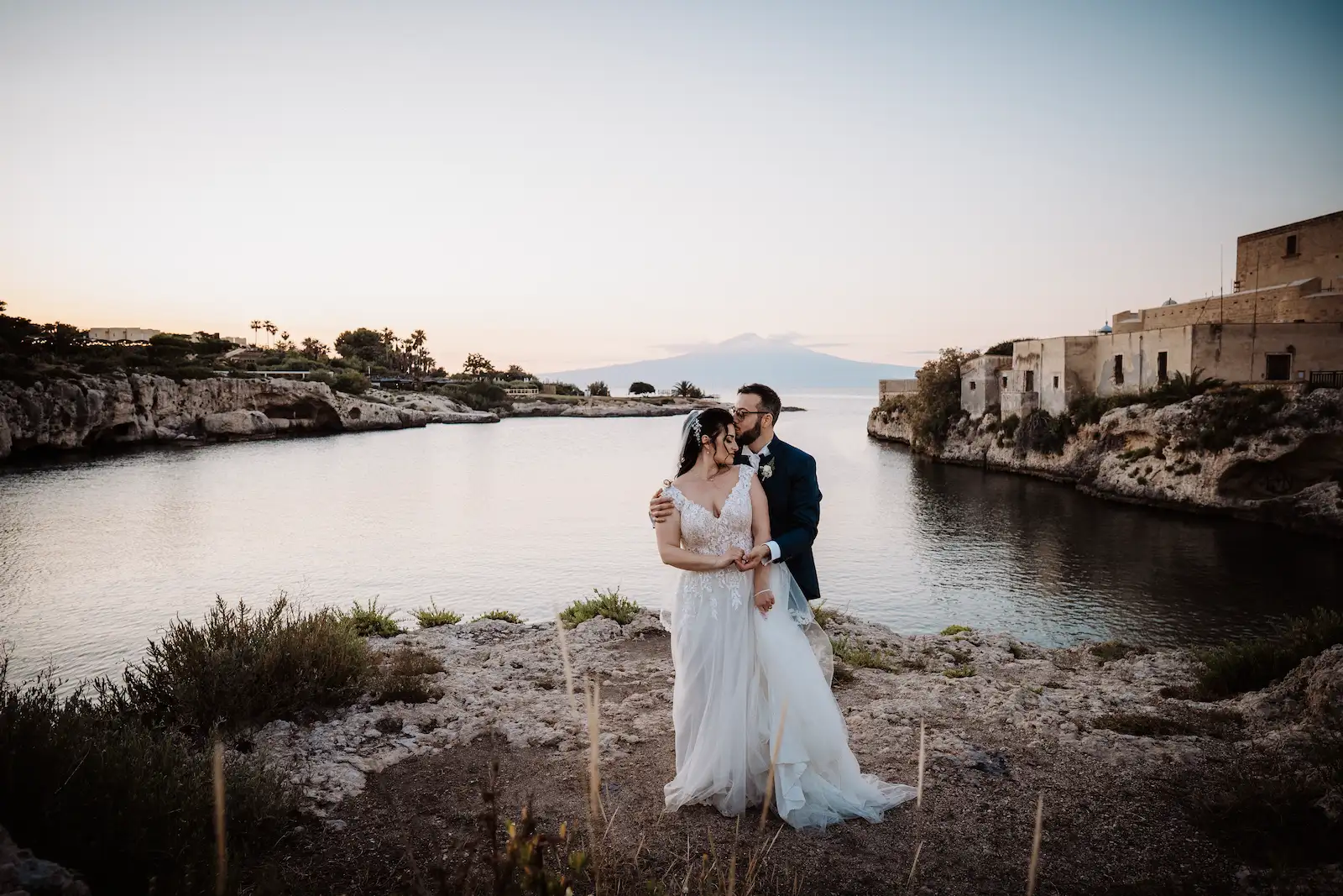 Foto di coppia di sposi che si abbracciano su una scogliera a Brucoli, Sicilia, con vista sulla baia e sul vulcano Etna in lontananza al tramonto
